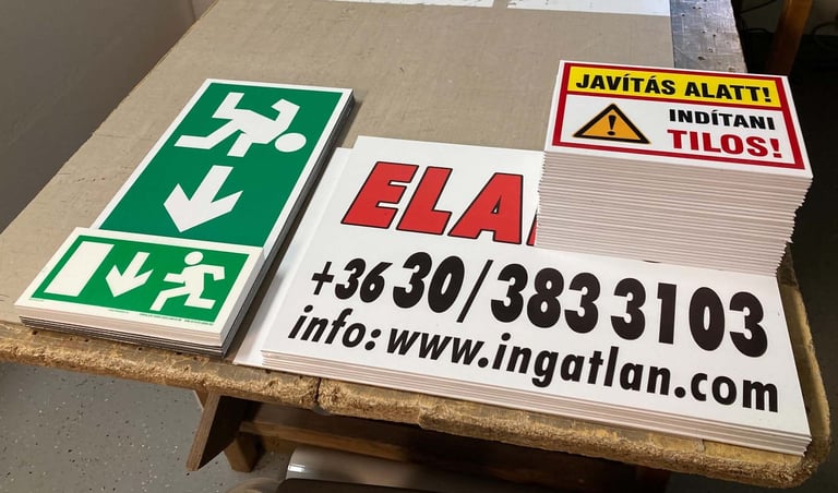 Collection of safety signs including green emergency exit sign, red ELA contact information sign, and yellow warning label on wooden shelf